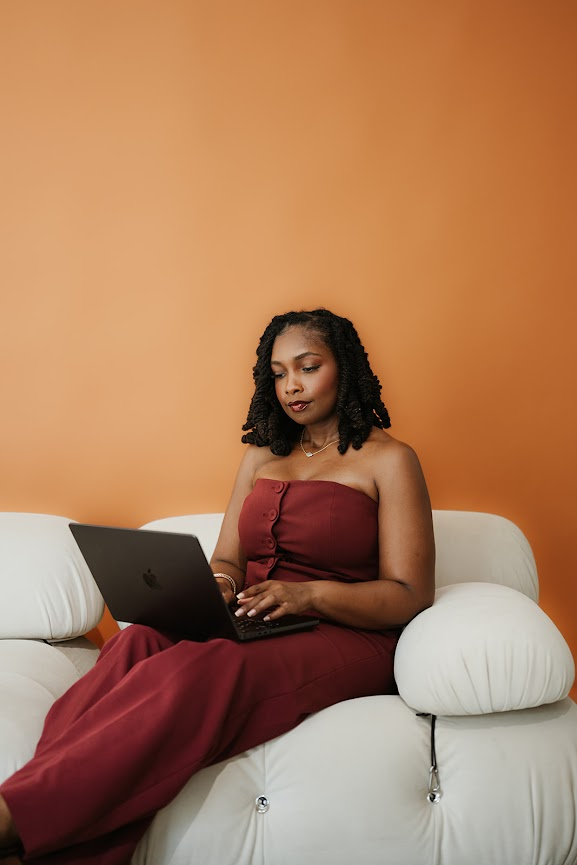 Kierra D., founder of The ROI Report, seated and working on a laptop in a burgundy jumpsuit
